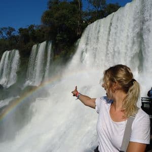 Una mujer rubia con cola de caballo señala un arcoíris que se forma en la neblina de una cascada grande y potente.