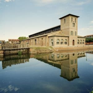 Un grande edificio in mattoni color sabbia con una torre si affaccia su un canale, il suo riflesso visibile nell'acqua calma.