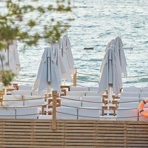 Ombrelloni bianchi chiusi e divani lounge vuoti su una terrazza in legno con vista mare.