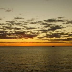 Due piccole barche in silhouette sul mare calmo durante un tramonto dorato sotto un cielo nuvoloso.