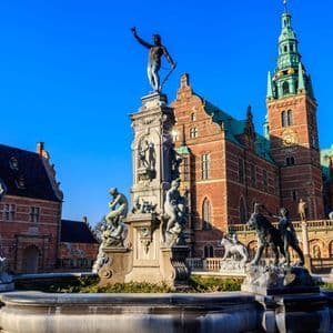 Une fontaine ornée de statues en bronze se trouve sur une place devant un bâtiment historique en briques rouges avec une tour horloge.