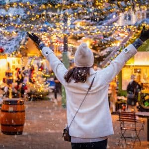 Una mujer vista de espaldas levanta los brazos en un mercado de Navidad iluminado mientras la nieve cae a su alrededor.