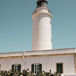 Un faro bianco con cupola in vetro si erge dietro un edificio con persiane verdi, con un muretto a secco e fichi d'India in primo piano.