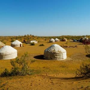 A camp of traditional yurts scattered across a sandy, arid landscape with low green shrubs under a clear blue sky.