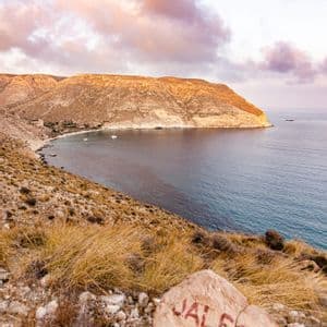 Una roca pintada con la palabra 'JALEO' se asienta en una colina seca y cubierta de hierba, con vistas a una cala con veleros y acantilados áridos al atardecer.