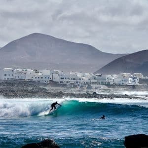 Un surfista cavalca una grande onda di fronte a una località costiera con edifici bianchi e colline vulcaniche sotto un cielo nuvoloso.
