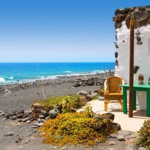 Un patio con un tavolo verde e due sedie di vimini all'esterno di una casa imbiancata su una spiaggia di sabbia nera accanto all'oceano.