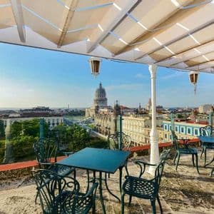 Una terrazza panoramica con tavoli e sedie in metallo verde si affaccia su un paesaggio urbano con un grande edificio a cupola sotto un cielo blu.