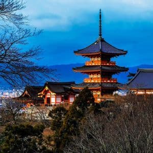 Una pagoda e un tempio giapponese arancioni a più livelli, su una collina che domina una città, incorniciati da alberi spogli sotto un cielo azzurro.