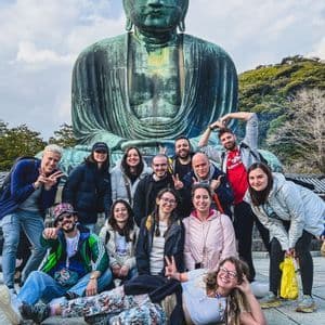Un gruppo WeRoad in viaggio posa e sorride per una foto davanti a una gigantesca statua bronzea del Buddha seduto sotto un cielo parzialmente nuvoloso.