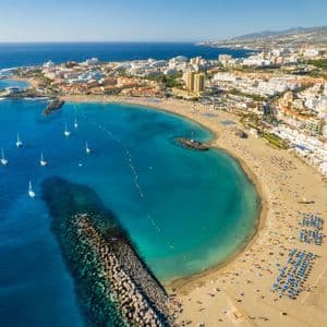 Luftaufnahme einer Küstenstadt mit einem belebten Sandstrand an einer türkisfarbenen Bucht voller Segelboote unter klarem blauem Himmel.