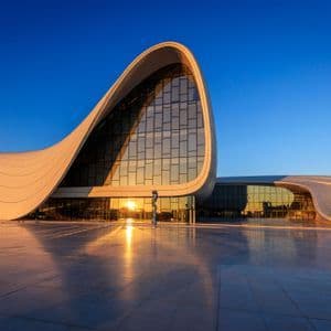 Un grande edificio bianco con un design moderno e curvo riflette la luce dorata del tramonto sulle sue finestre di vetro e sulla piazza sottostante.