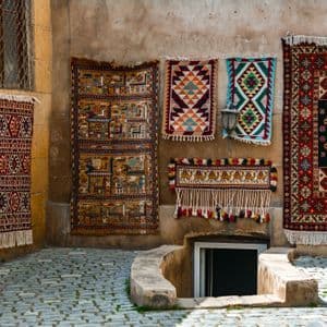 Una varietà di tappeti tradizionali a fantasia è appesa a un muro di pietra logorato lungo una strada acciottolata.