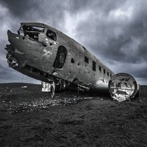 Das Wrack eines Flugzeugrumpfes liegt auf einem Feld aus schwarzem Sand unter einem stark bewölkten Himmel.