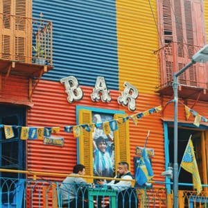 Dos hombres sentados en una mesa en un balcón frente a un edificio colorido de chapa con un letrero de 'BAR'.