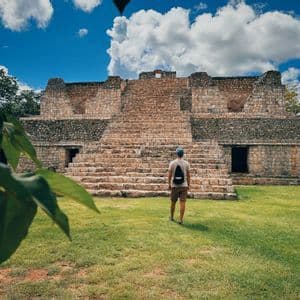 Un uomo visto di spalle, in piedi su un prato verde e che guarda una grande antica piramide di pietra sotto un cielo blu con nuvole.