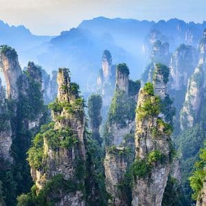 Alte formazioni rocciose a pilastro coperte di vegetazione verde si ergono da una valle nebbiosa con montagne sullo sfondo.