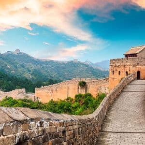 Un antico muro di pietra con una torre di guardia si snoda attraverso verdi montagne sotto un cielo colorato al tramonto.