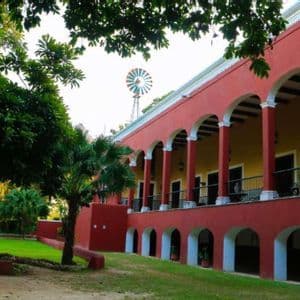 Una hacienda rossa a due piani con una veranda ad arco e un mulino a vento sul tetto si erge accanto a un prato rigoglioso con palme.