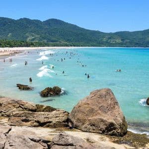 Una spiaggia affollata in una giornata di sole con persone che nuotano in acque turchesi, con montagne verdi e lussureggianti sullo sfondo.