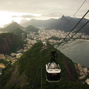 Una funivia scende su una città costiera incastonata tra verdi montagne, con una baia piena di barche e una statua su una cima lontana.