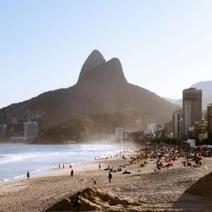 Un'ampia veduta di una spiaggia sabbiosa affollata vicino a una città, con grandi montagne verdi che si ergono sullo sfondo sotto un cielo fosco.