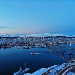 Vista aérea de una ciudad costera nevada al anochecer, con un puente largo sobre el agua y montañas al fondo.