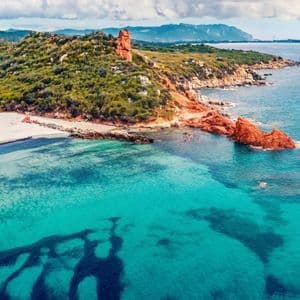 Vista aerea di una costa con acqua turchese, una spiaggia di sabbia bianca, rocce rosse e colline verdi sotto un cielo nuvoloso.