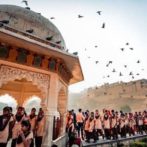 Un gruppo di studentesse in uniforme passa davanti a un padiglione decorato, con uccelli che volano in alto e un grande forte sullo sfondo.