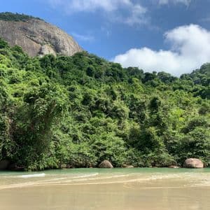 Una spiaggia sabbiosa con acqua calma e verde chiaro, ai piedi di una montagna lussureggiante ricoperta di giungla, sotto un cielo azzurro.