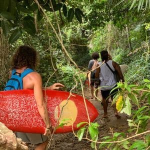 Un viaje en grupo de WeRoad caminando en fila india con tablas de surf por un estrecho sendero de tierra a través de una exuberante selva verde.