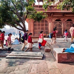 Un gruppo di scolare in divisa attraversa un cortile in pietra, passando accanto a un uomo che riposa su una pedana.