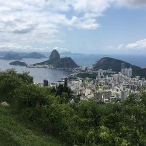 Vista elevada desde una exuberante colina verde, con vistas a una ciudad costera, una bahía y una prominente montaña cónica bajo un cielo nublado.