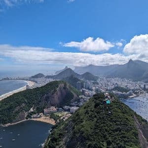 Vista aérea de una ciudad costera con playas y exuberantes montañas verdes que bordean el océano bajo un cielo azul y nublado.