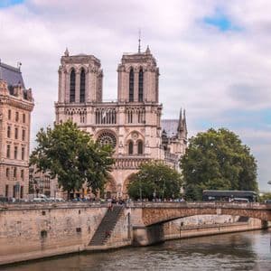 Una gran catedral gótica con torres gemelas se alza detrás de un puente de piedra que cruza un río en una ciudad bajo un cielo nublado.