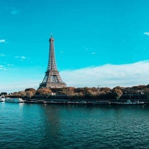 La Torre Eiffel si erge di fronte a un ampio fiume fiancheggiato da alberi autunnali e barche ormeggiate, sotto un cielo azzurro brillante.