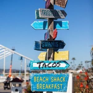 Un cartello in legno colorato con diverse insegne dipinte a mano che indicano spiagge e punti di ristoro, su uno sfondo di cielo azzurro.