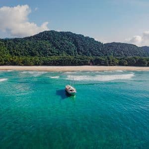 Luftaufnahme eines weißen Segelboots, das in türkisfarbenem Wasser ankert, mit Sandstrand und üppigem, grünem Berg im Hintergrund.