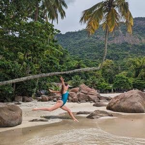 Eine Frau in blauem Badeanzug springt mitten in der Luft an einem Sandstrand mit großen Felsen, Palmen und einem bewaldeten Berg im Hintergrund.