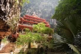 Un temple rouge traditionnel à plusieurs niveaux, intégré à une falaise rocheuse et entouré d'une végétation luxuriante de jungle.