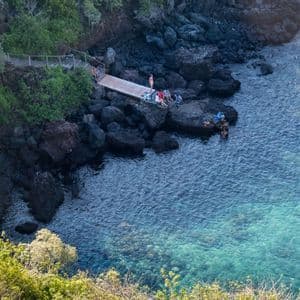 Luftaufnahme einer WeRoad-Gruppe, die auf einem Holzsteg entspannt und im klaren, blauen Meer vor einer felsigen Küste schwimmt.