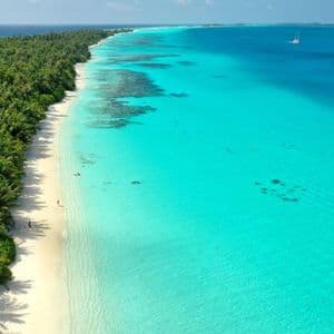 Una vista aerea di una lunga isola tropicale con una spiaggia di sabbia bianca, palme e acqua cristallina turchese con una barca a vela in lontananza.