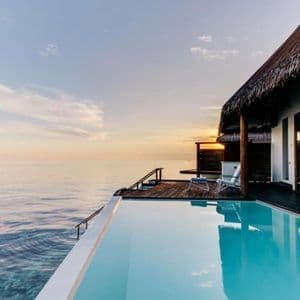 Un bungalow sull'acqua con tetto di paglia e piscina a sfioro si affaccia sull'oceano al tramonto.