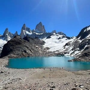 Un lago alpino turchese circondato da una riva rocciosa e montagne frastagliate e innevate sotto un cielo limpido e luminoso.