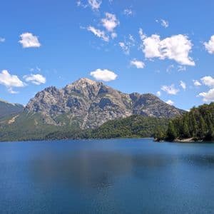 Una grande montagna rocciosa con pendii boscosi si erge sopra un lago blu calmo sotto un cielo parzialmente nuvoloso.