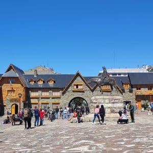 Una statua equestre si trova in una vivace piazza cittadina piena di gente, di fronte a un edificio in pietra e legno, sotto un cielo azzurro chiaro.