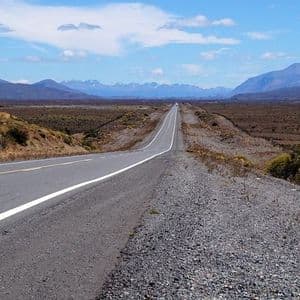 Una strada diritta e asfaltata si estende attraverso una vasta pianura arida verso una catena montuosa lontana sotto un cielo parzialmente nuvoloso.