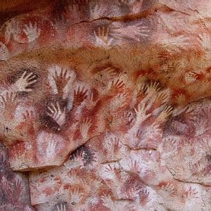 Antiche pitture rupestri con numerosi stencil di mani in pigmenti rossi, bianchi e scuri su una parete rocciosa grezza.