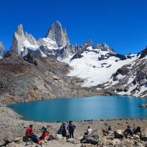Un viaggio di gruppo WeRoad si rilassa sulla riva rocciosa di un lago alpino turchese, ai piedi di montagne frastagliate e innevate.