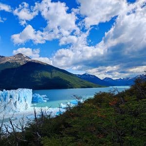 Un'immensa parete glaciale incontra un lago turchese pieno di iceberg, con montagne verdi sullo sfondo sotto un cielo azzurro nuvoloso.